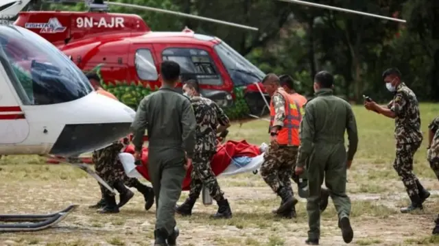 Army personnel carry the wounded on a stretcher covered with a red coat. The front of a white helicopter appears, with a second red helicopter behind.