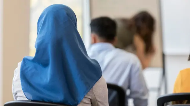 From behind, an unknown woman in a hijab is sitting on a chair in a classroom. Other colleagues are sitting in front of him.
