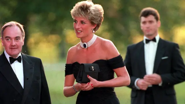 Princess Diana has short blonde hair. She wears a black off-the-shoulder dress with a necklace with white pearls and a large black bead in the middle. Next to him is British businessman Peter Palmeau, wearing a black formal suit with a white shirt and black bow tie. Behind him, on the other side, stands a man also dressed in official clothes, but his features are indistinct.